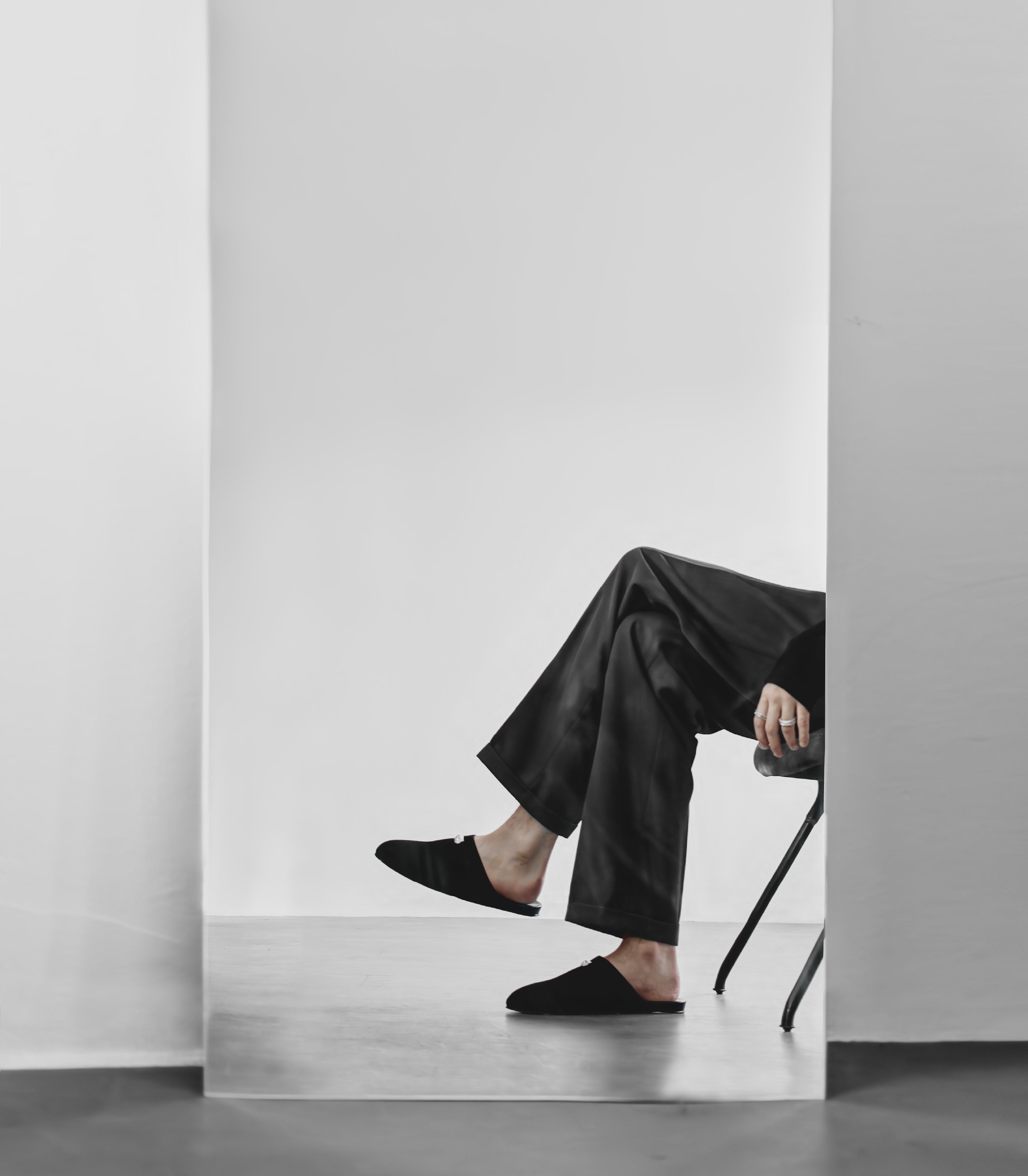 Man wearing black suede slippers and pants sitting on a chair half infront of a mirror.