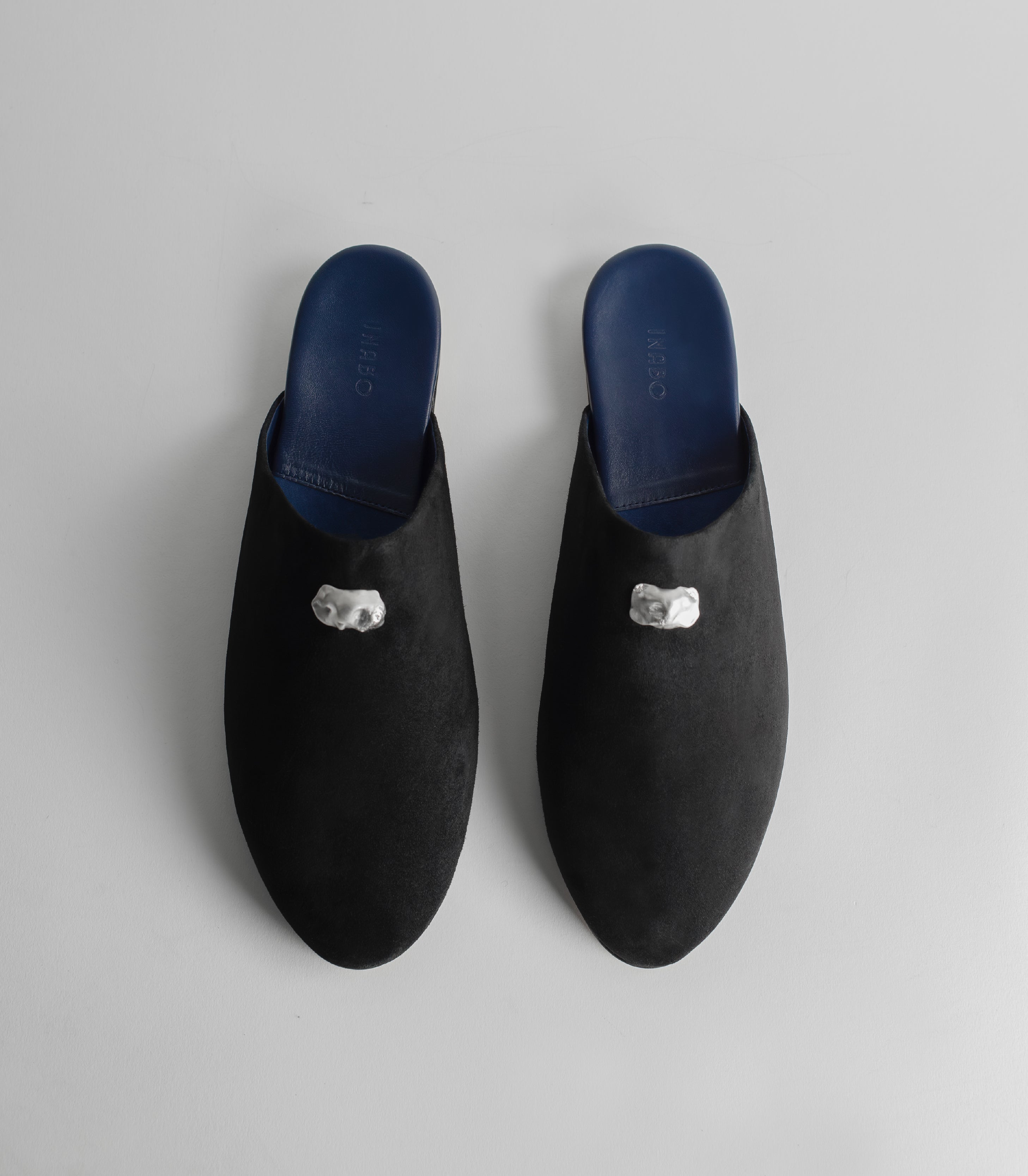 A pair of black suede slippers with a silver jewelry attached on a gray background.