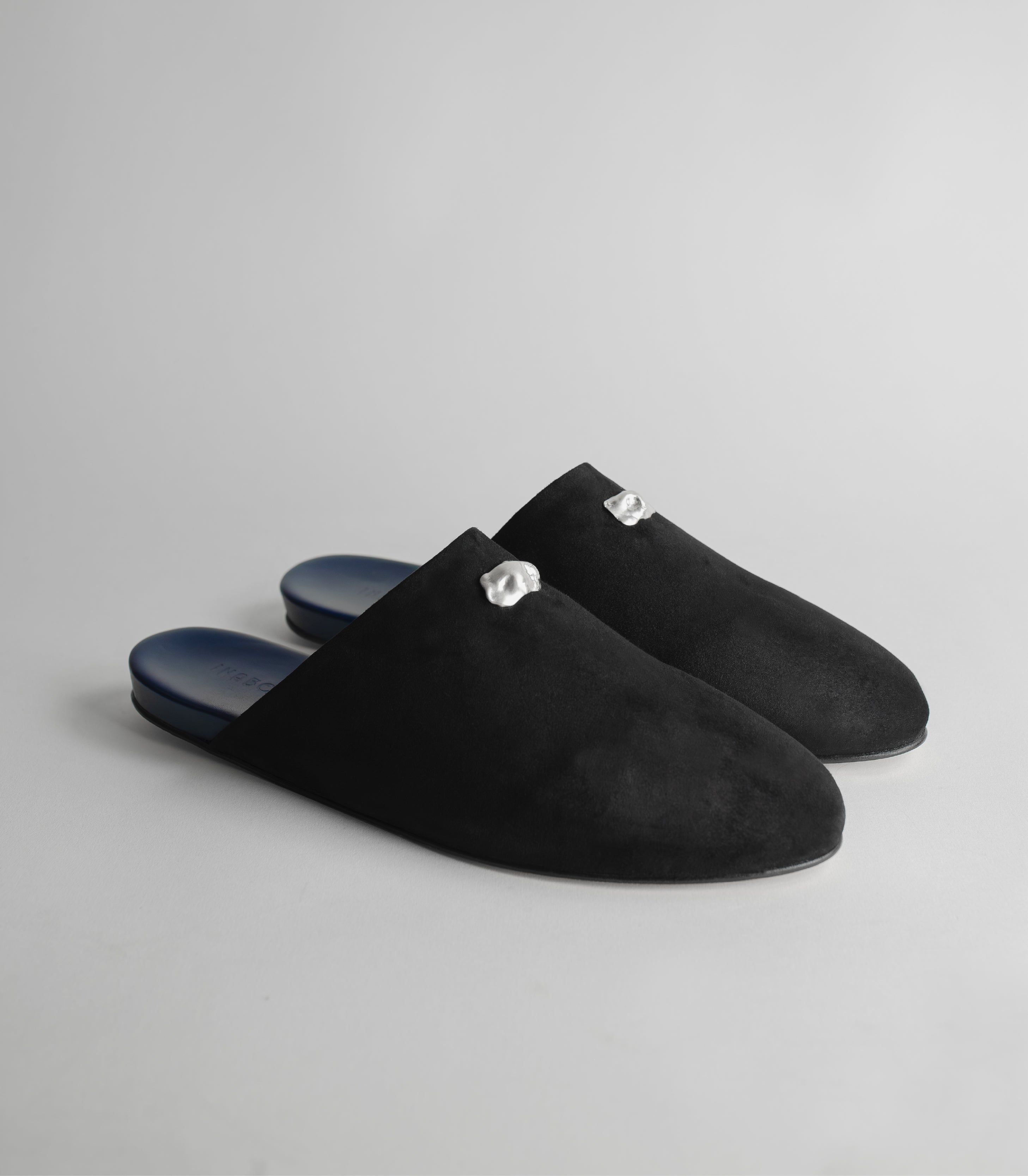 Black suede slippers with silver buckles on a gray background.
