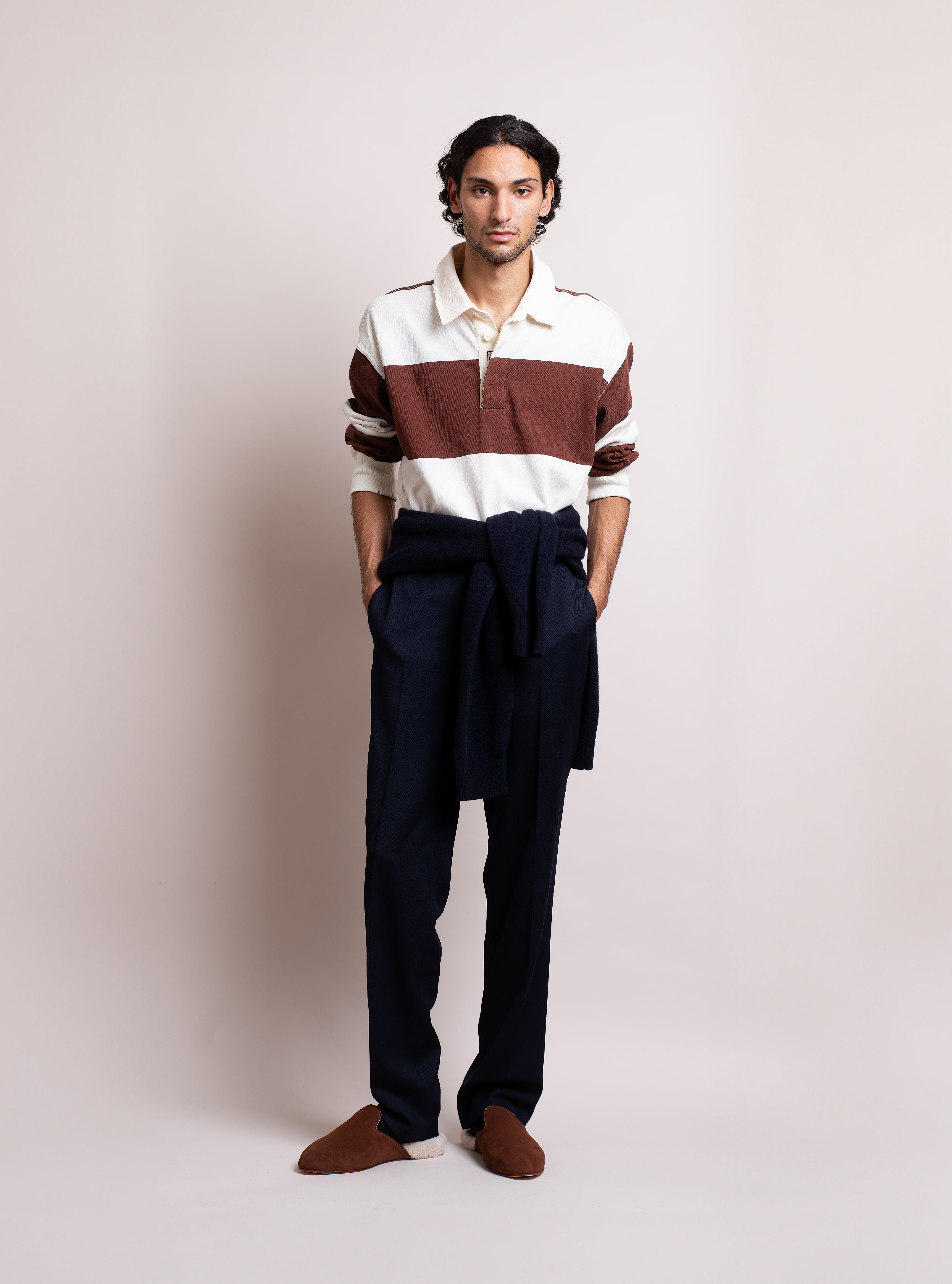 Man wearing a striped shirt, dark navy pants, and tobacco brown suede slippers with white shearling against a plain background.