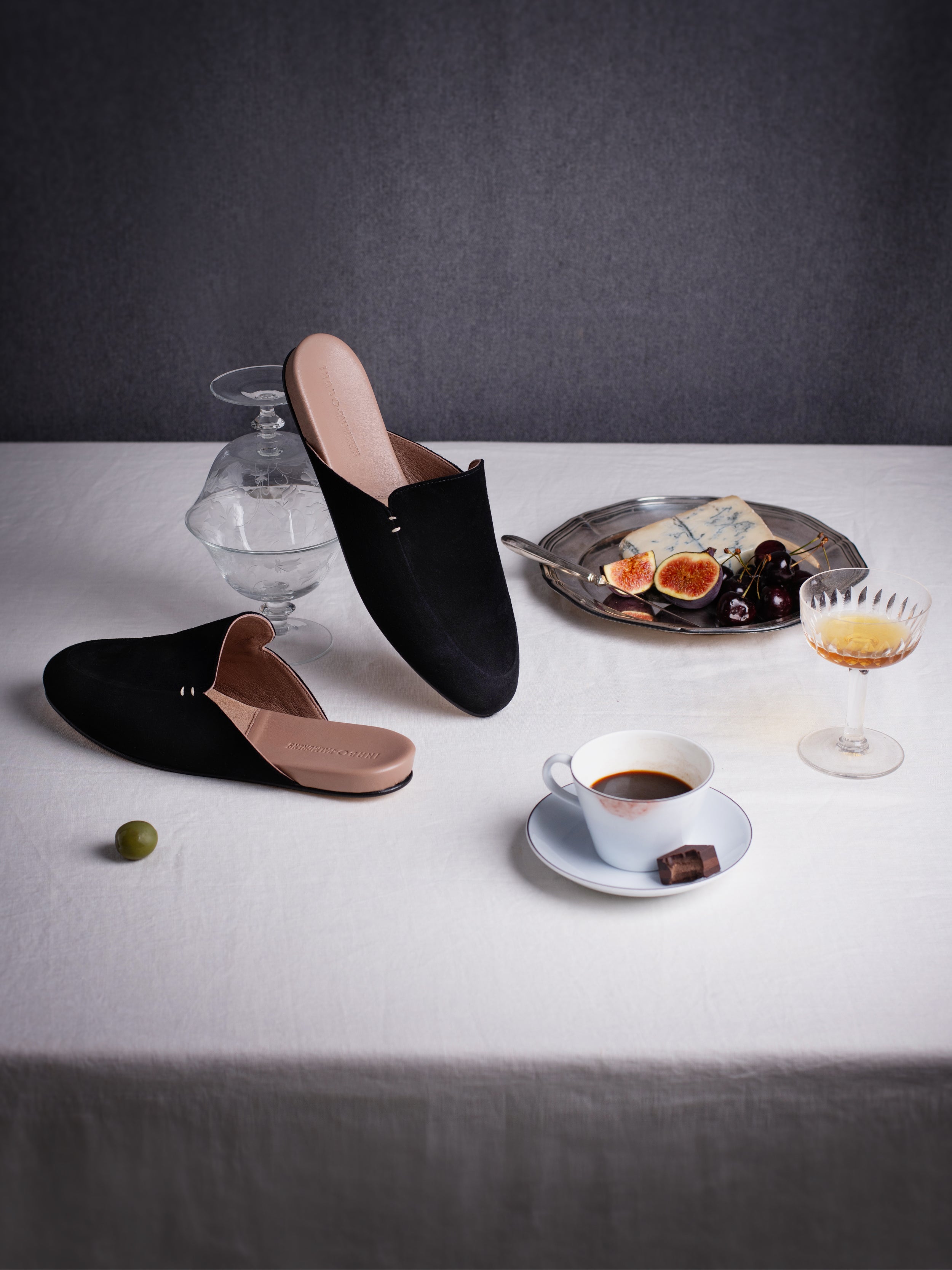 A still life image with a pair of black suede slippers, a cup of coffee, a drink and a plate with cheese.