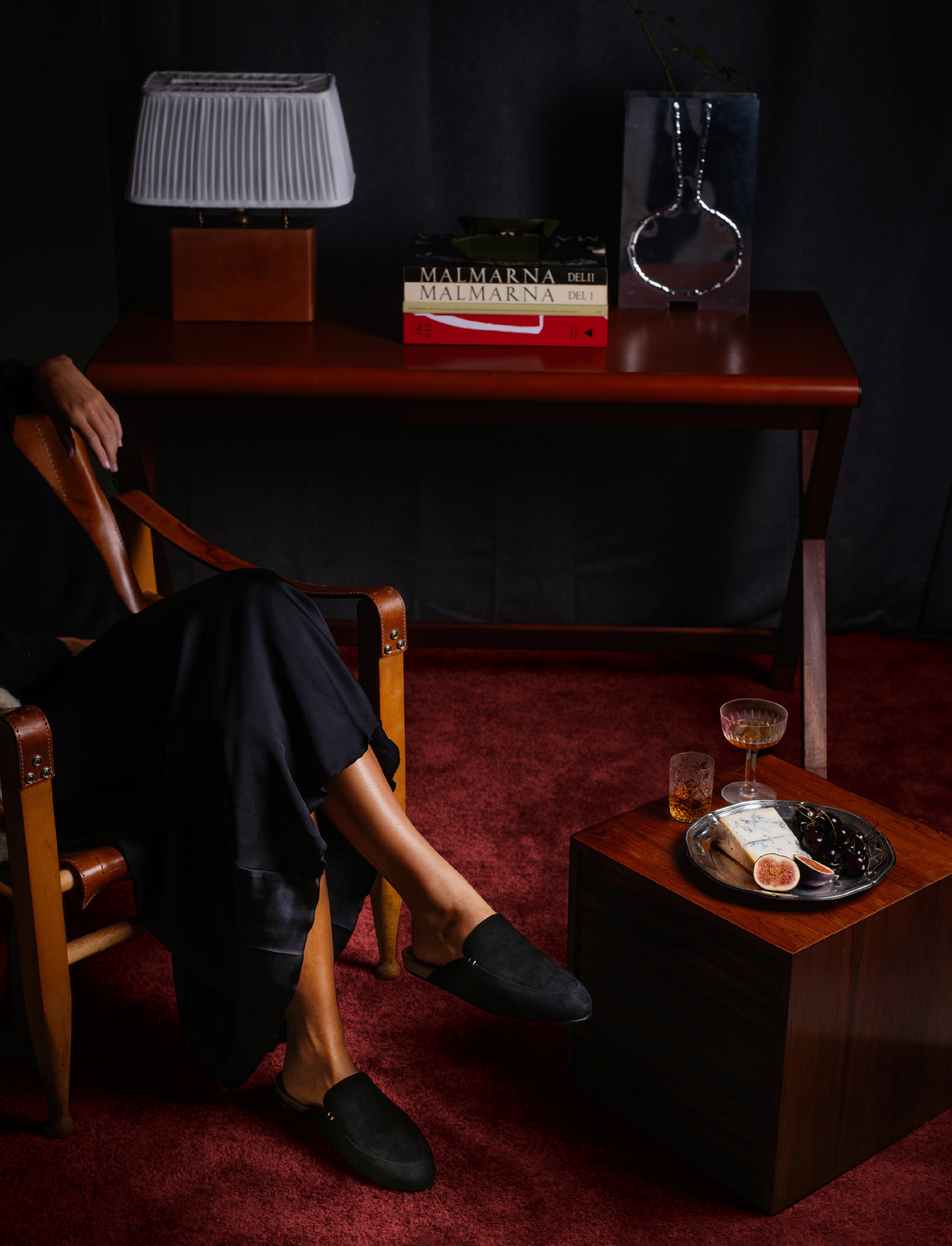 Woman sitting in a chair with a table displaying books and a glass of wine in a dimly lit room, wearing a pair of black suede slippers and a black silk skirt.