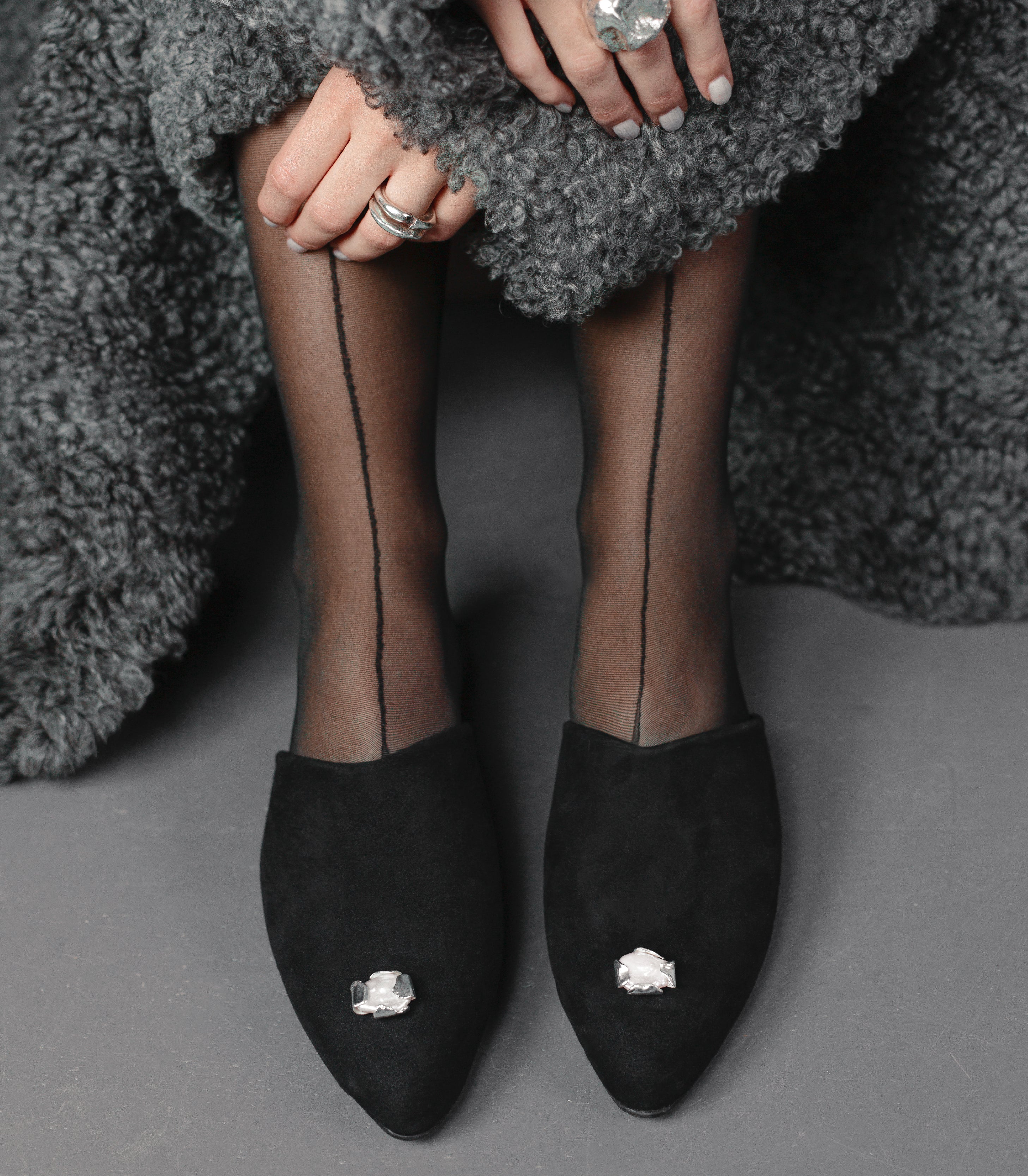 Black pointed-toe house shoes with handcrafted silver jewelry worn by a woman in a gray shearling coat.