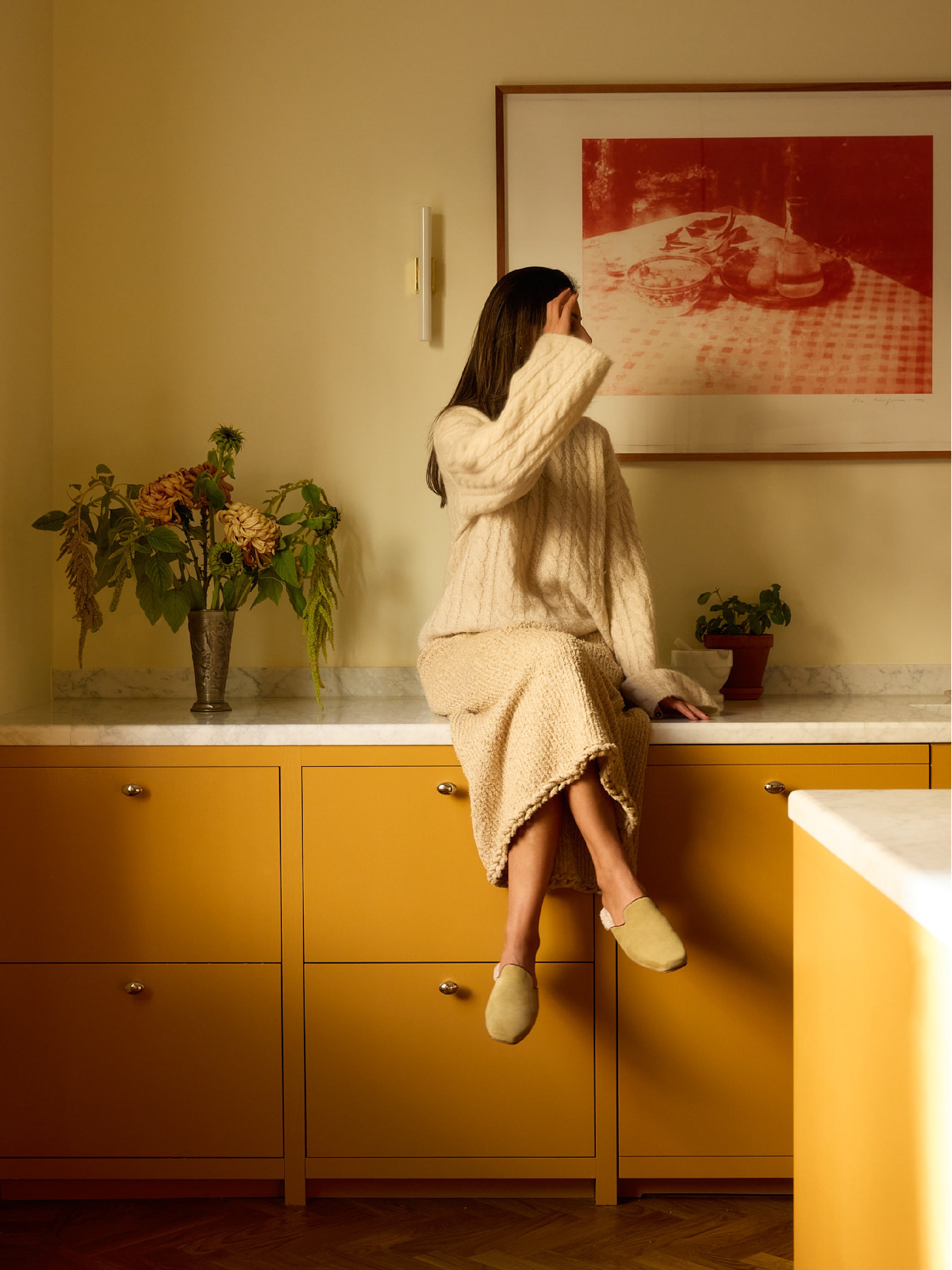 Woman sitting on a kitchen counter with yellow cabinets and a red painting on the wall, wearing a pair of olive green suede slippers with white shearling lining.