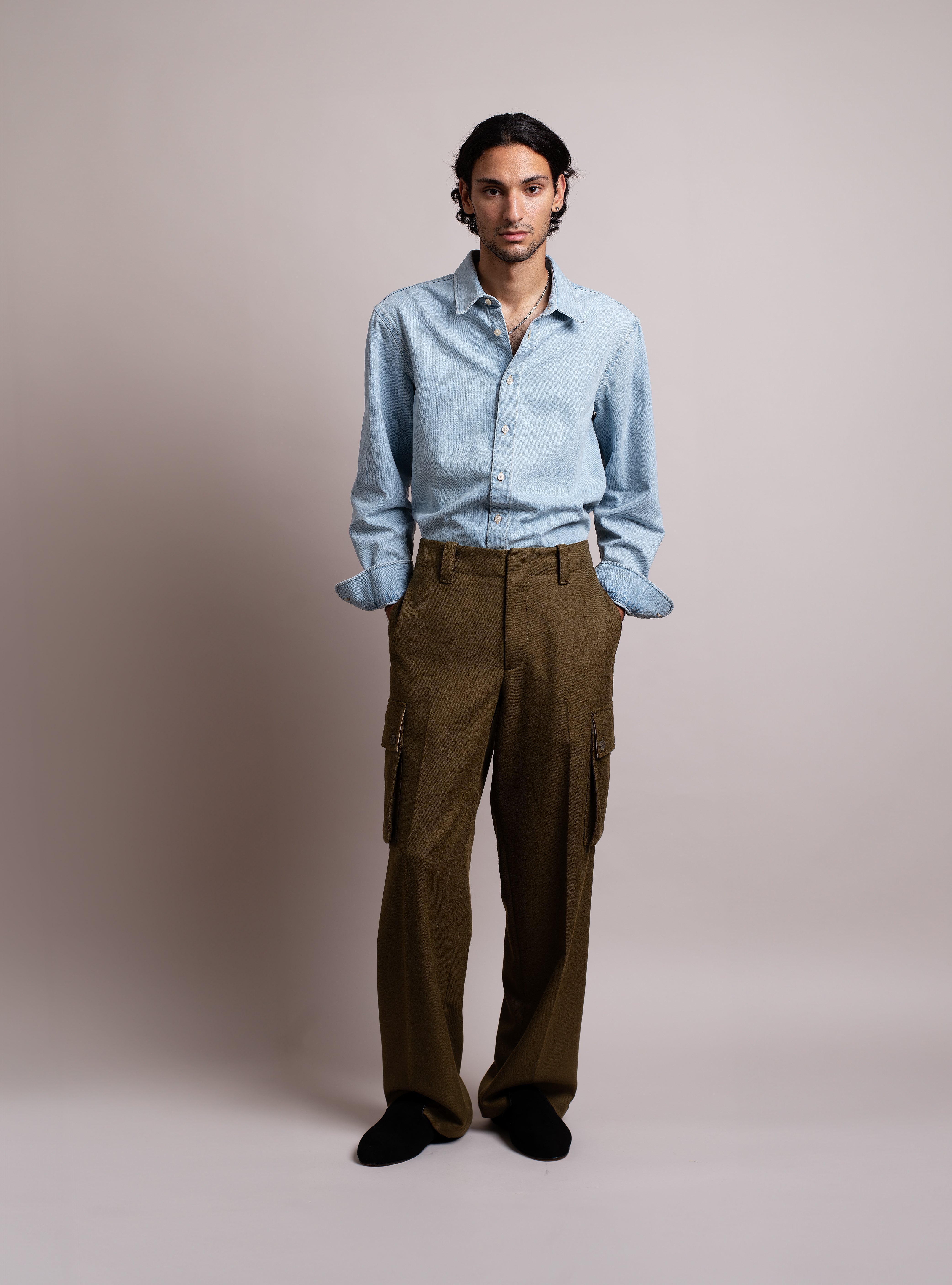 Man wearing a light blue jeans shirt, khaki green cargo pants, and black suede slippers with black shearling against a plain background.
