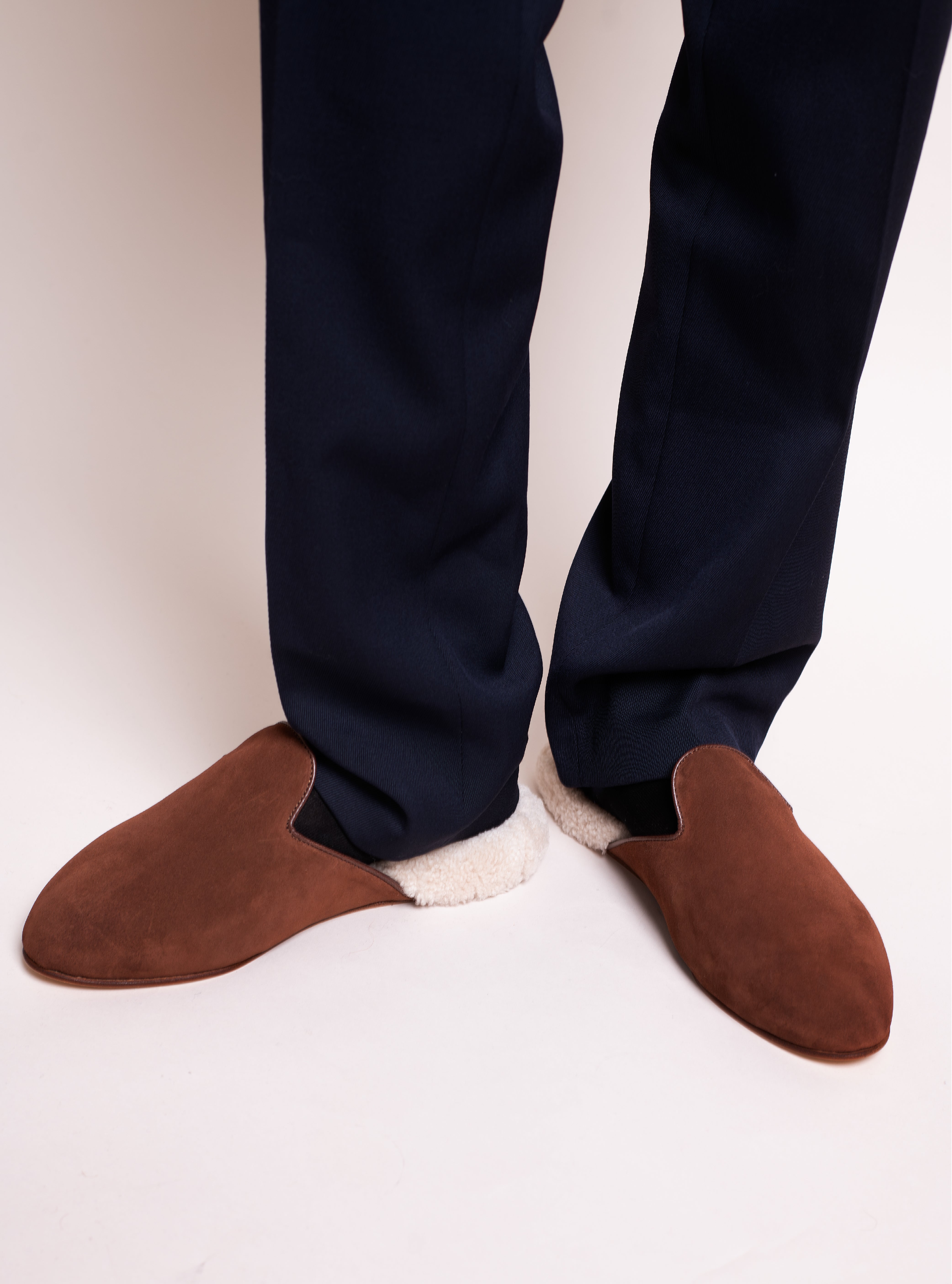 Man wearing tobacco brown suede slippers with white shearling, paired with navy blue pants.