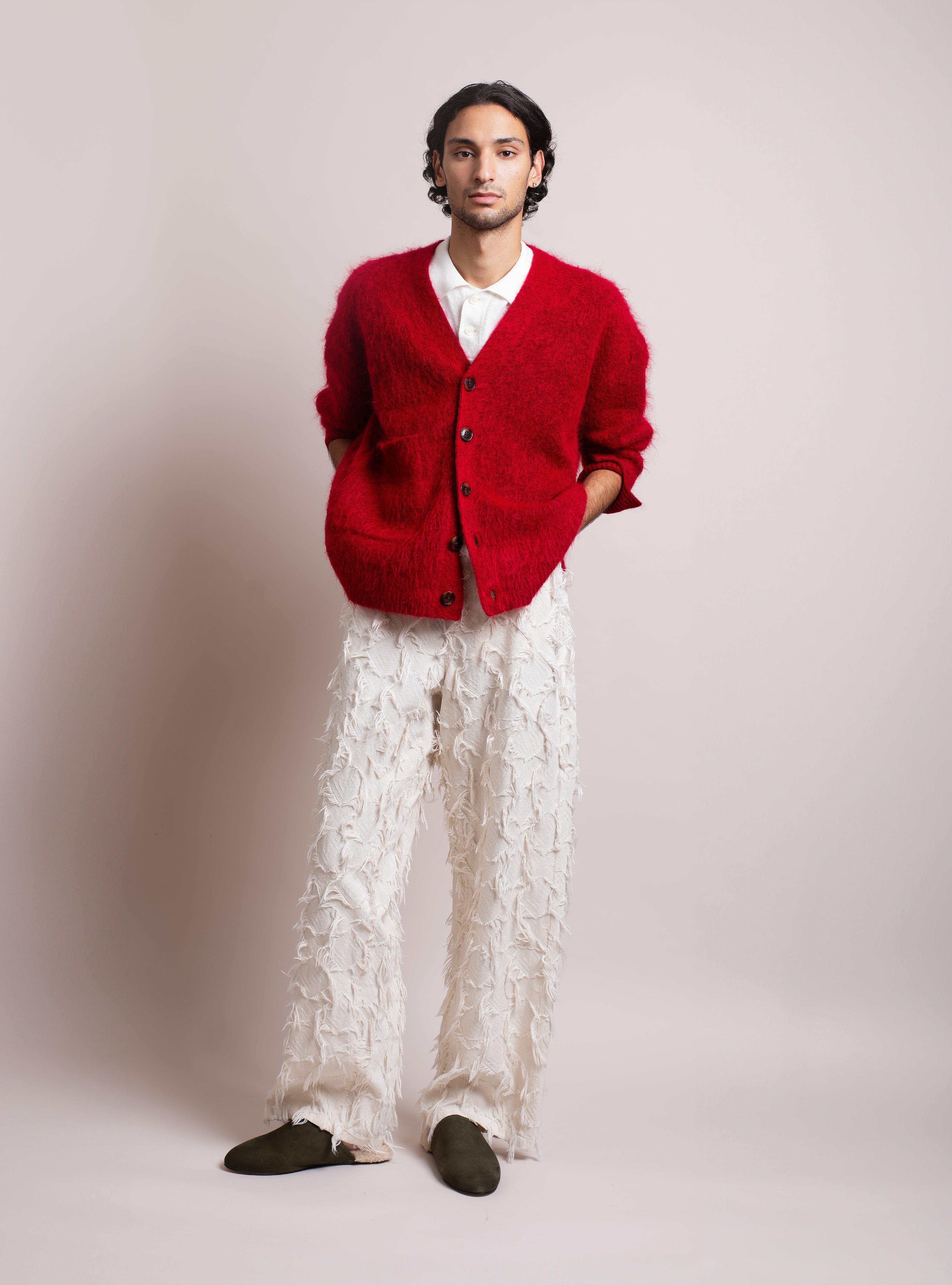 Man wearing a red cardigan over a white shirt, white pants, and army green suede slippers with white shearling on a plain background.