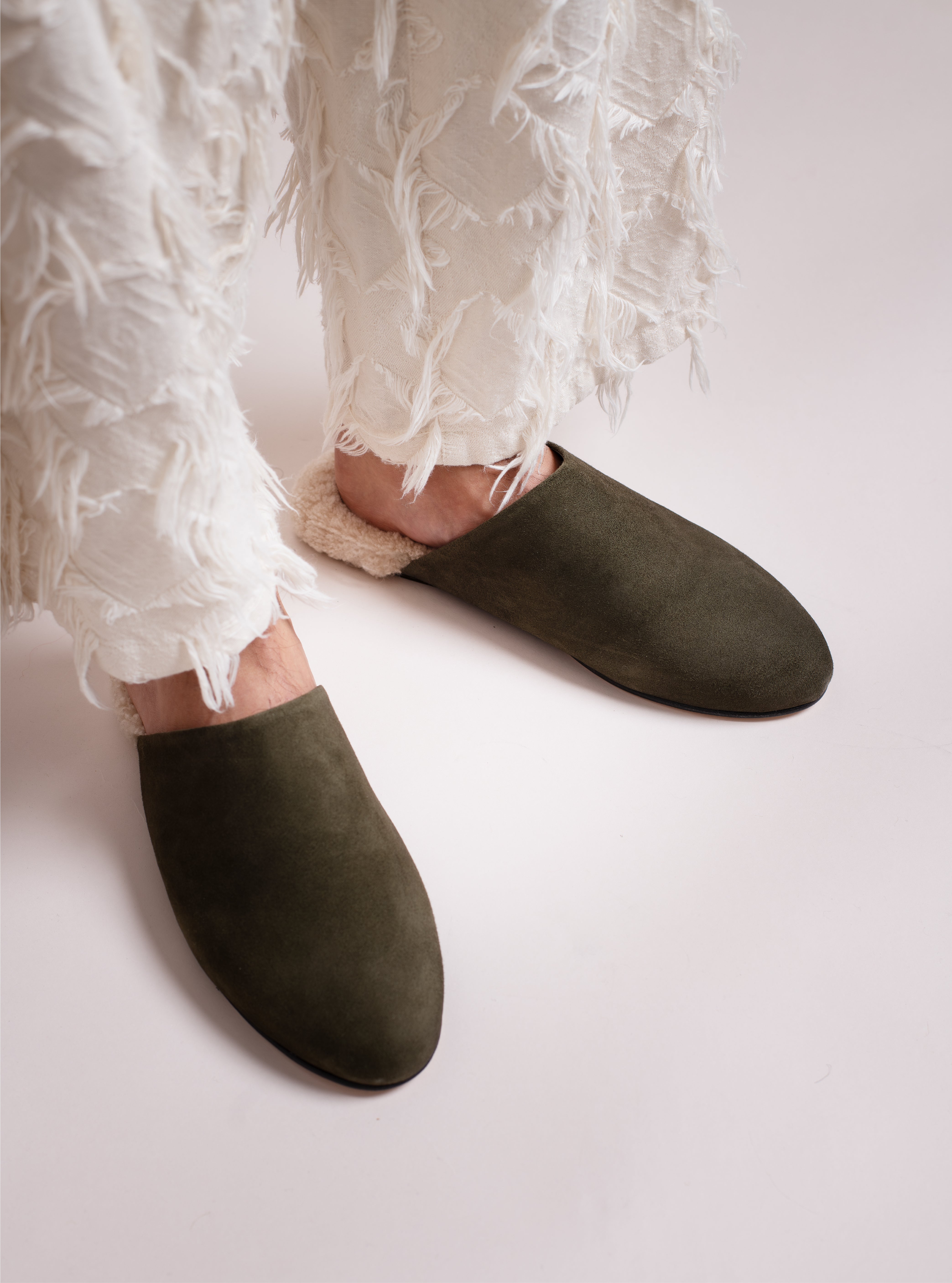 Green suede slippers with white shearling worn with white pants on a light background