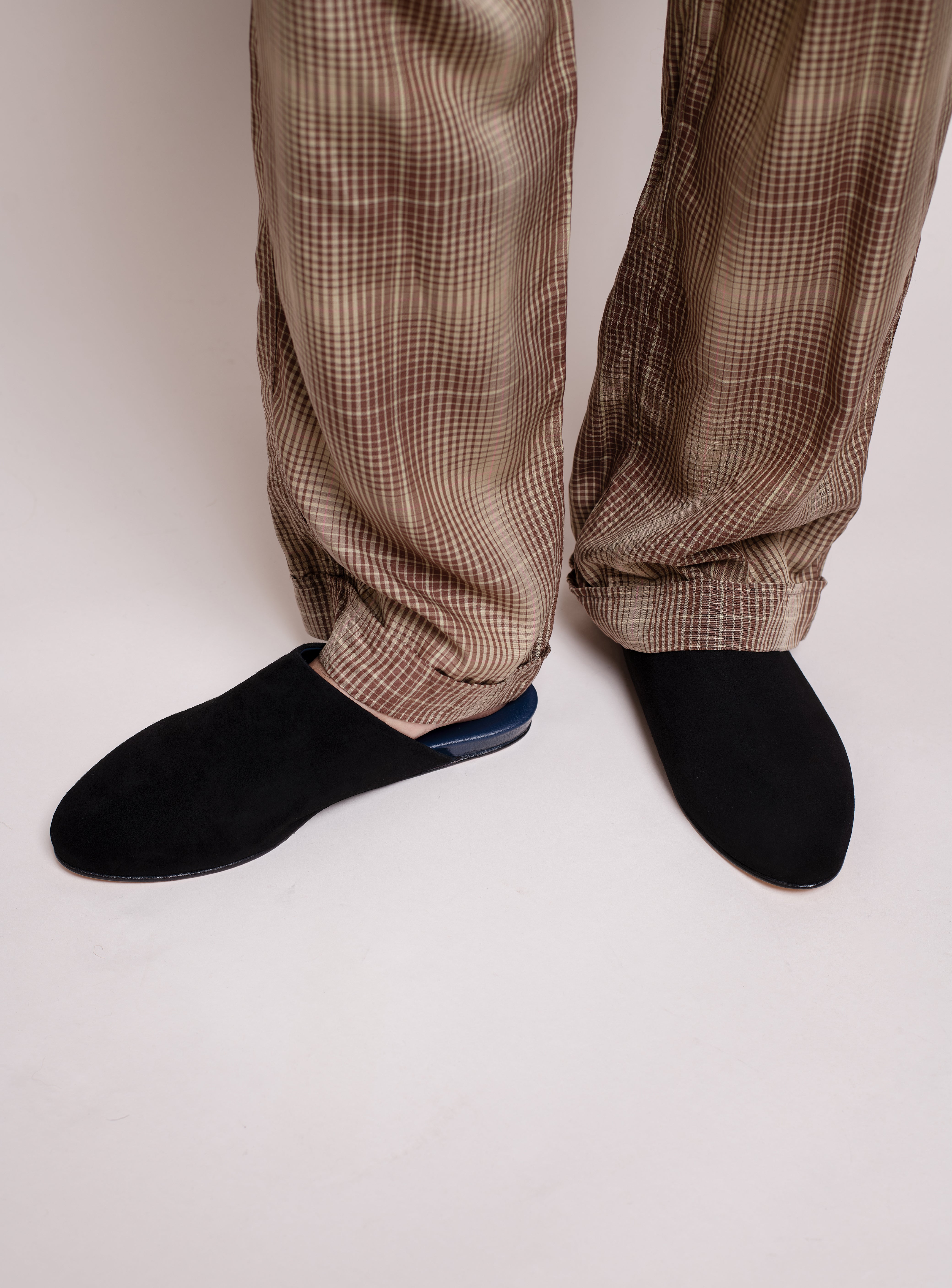 Man wearing plaid pants and black suede house slippers on a plain background.