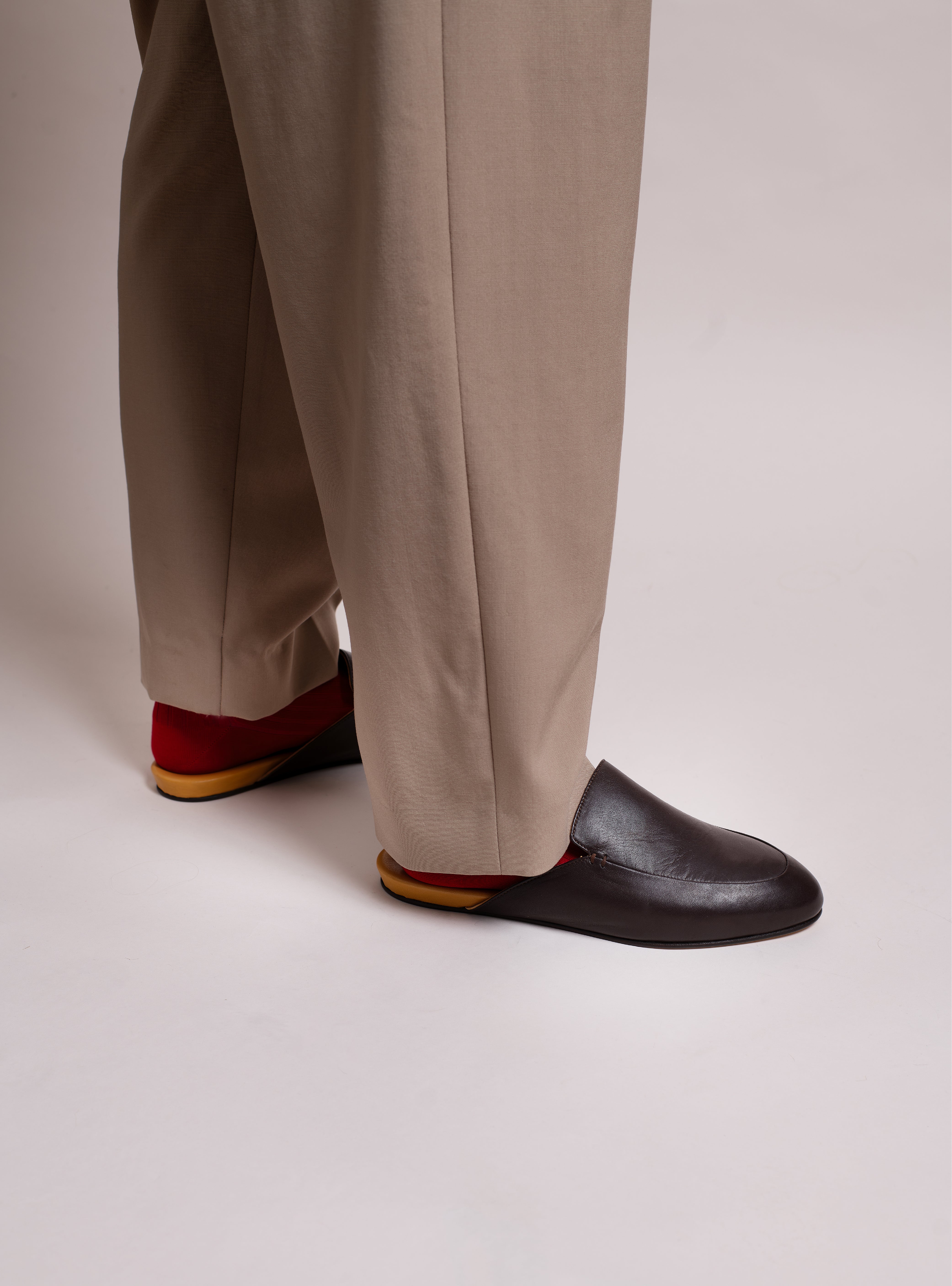 Man wearing beige pants and dark brown leather slippers with yellow soles on a plain background