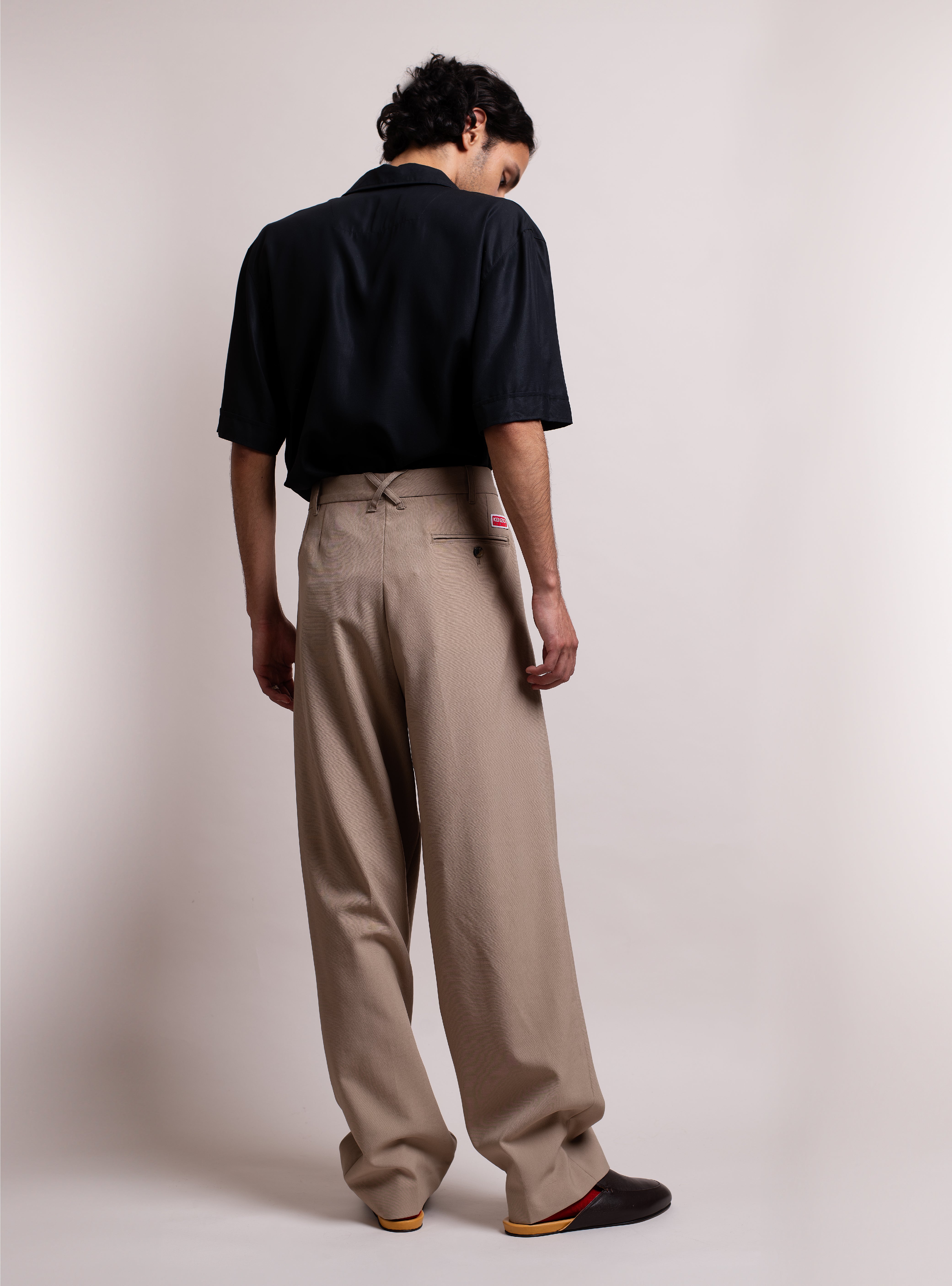 Man wearing a black shirt, beige pants, and dark brown leather slippers with a mustard yellow footbed against a plain background.