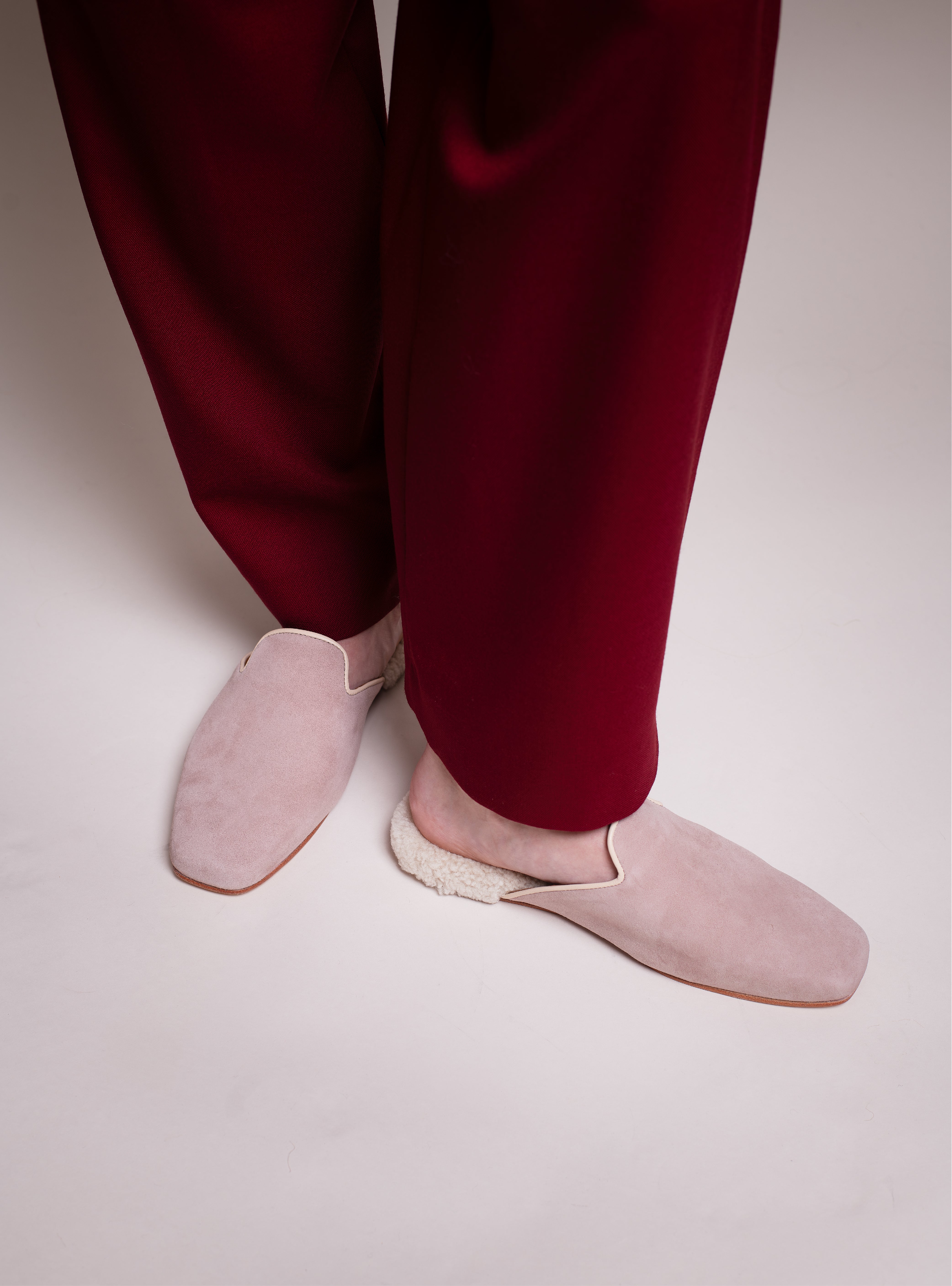 Women's light pink suede slippers with white shearling worn with red pants on a light background