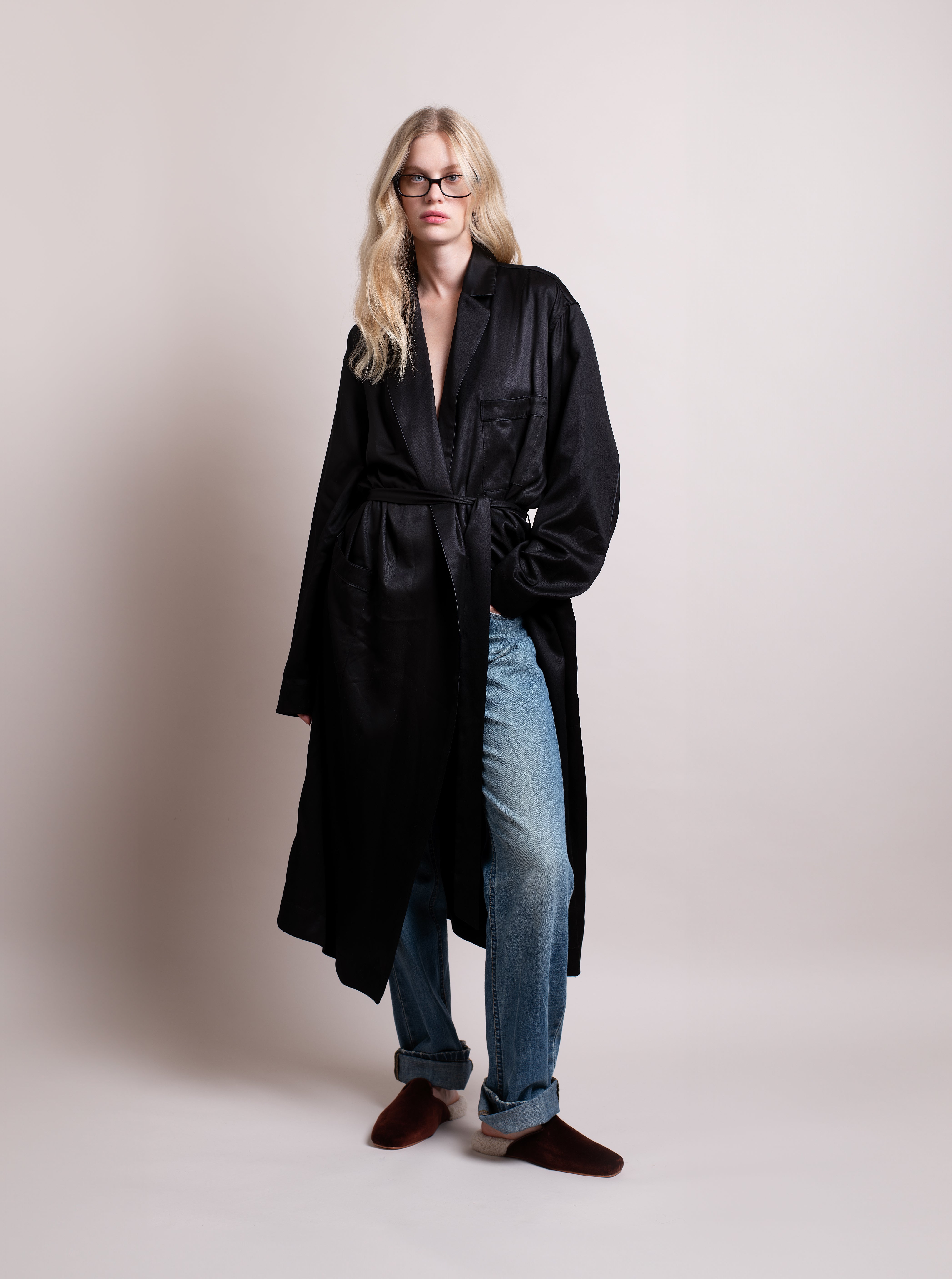 Woman wearing a black satin robe, blue jeans, and brown velvet slippers with white shearling against a plain background.