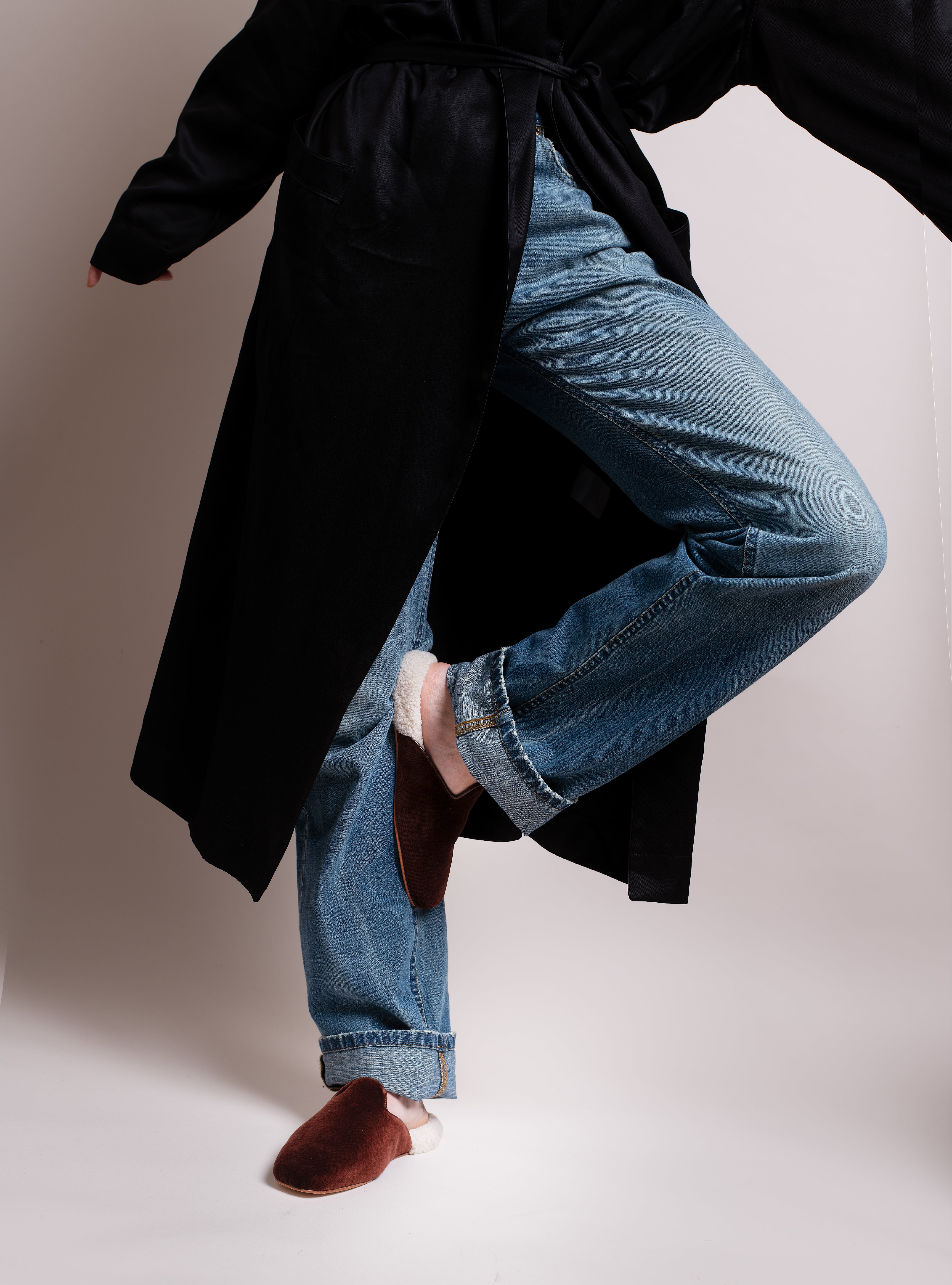 Woman wearing a black silk robe, blue jeans, and brown velvet slippers with white shearling on a plain background.