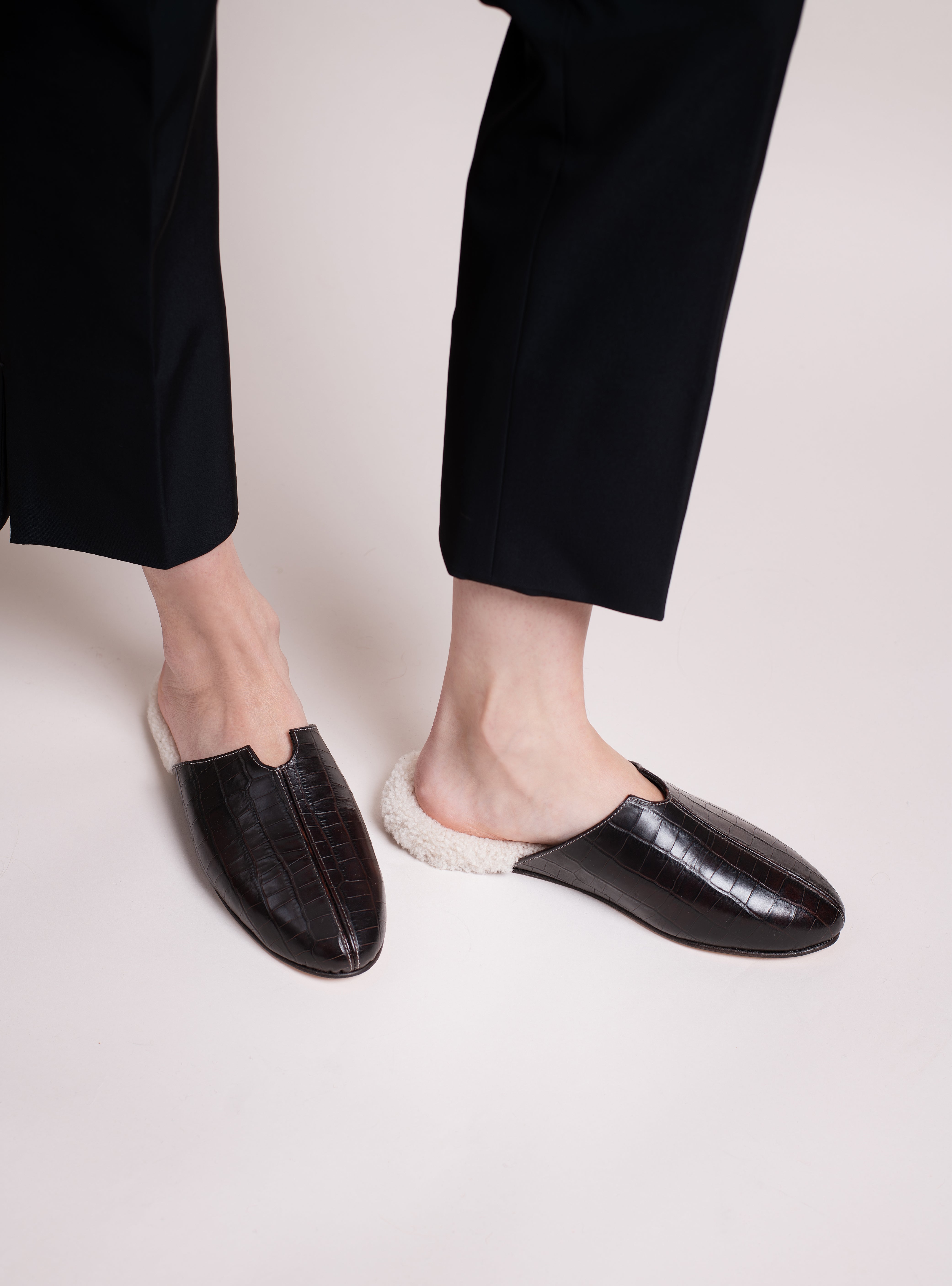 Women's croco-embossed, dark brown leather slippers worn with black pants on a light background