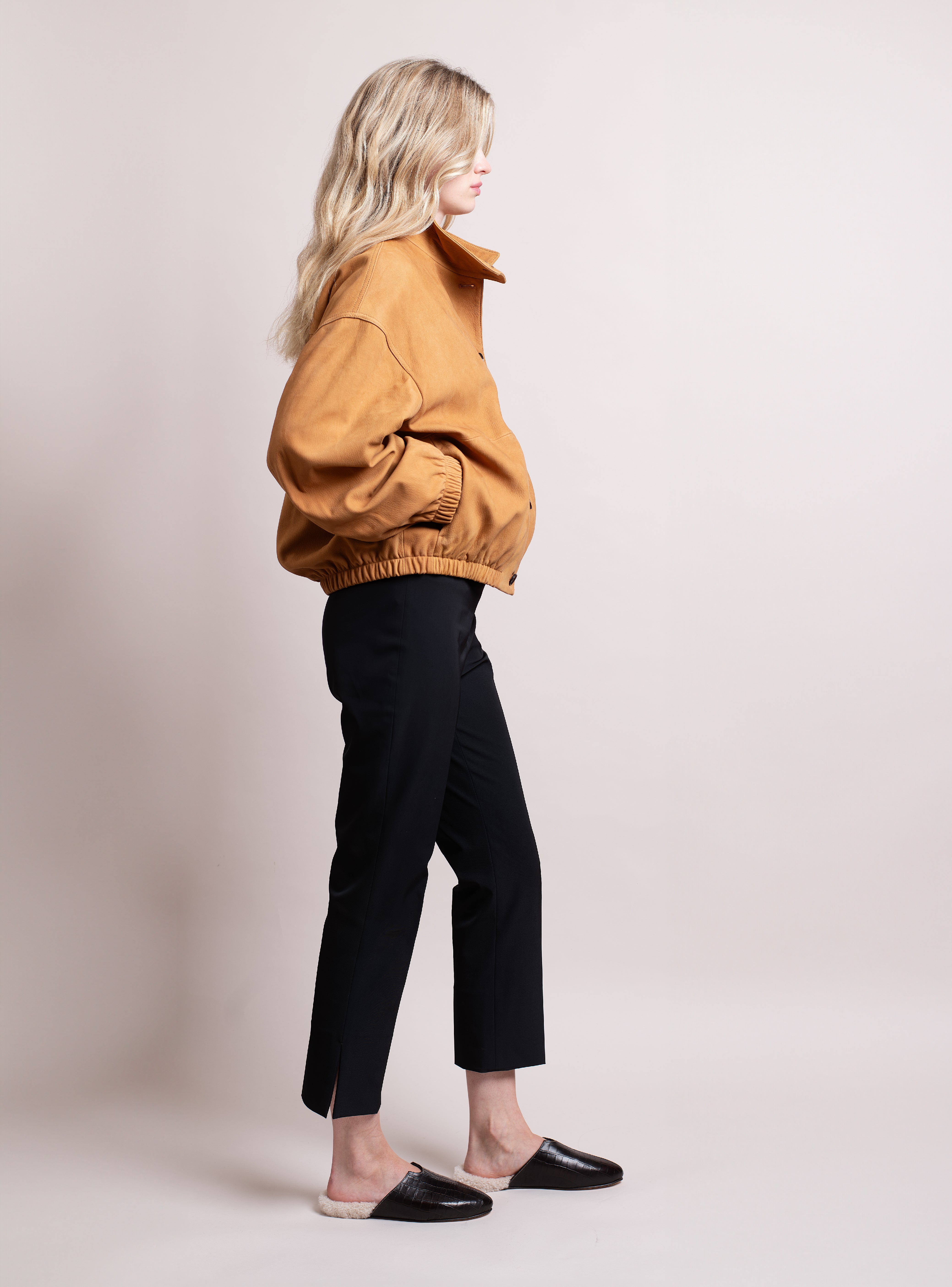 Woman wearing a cognac suede jacket, black pants, and croc-embossed slippers in dark brown and white shearling on a plain background.