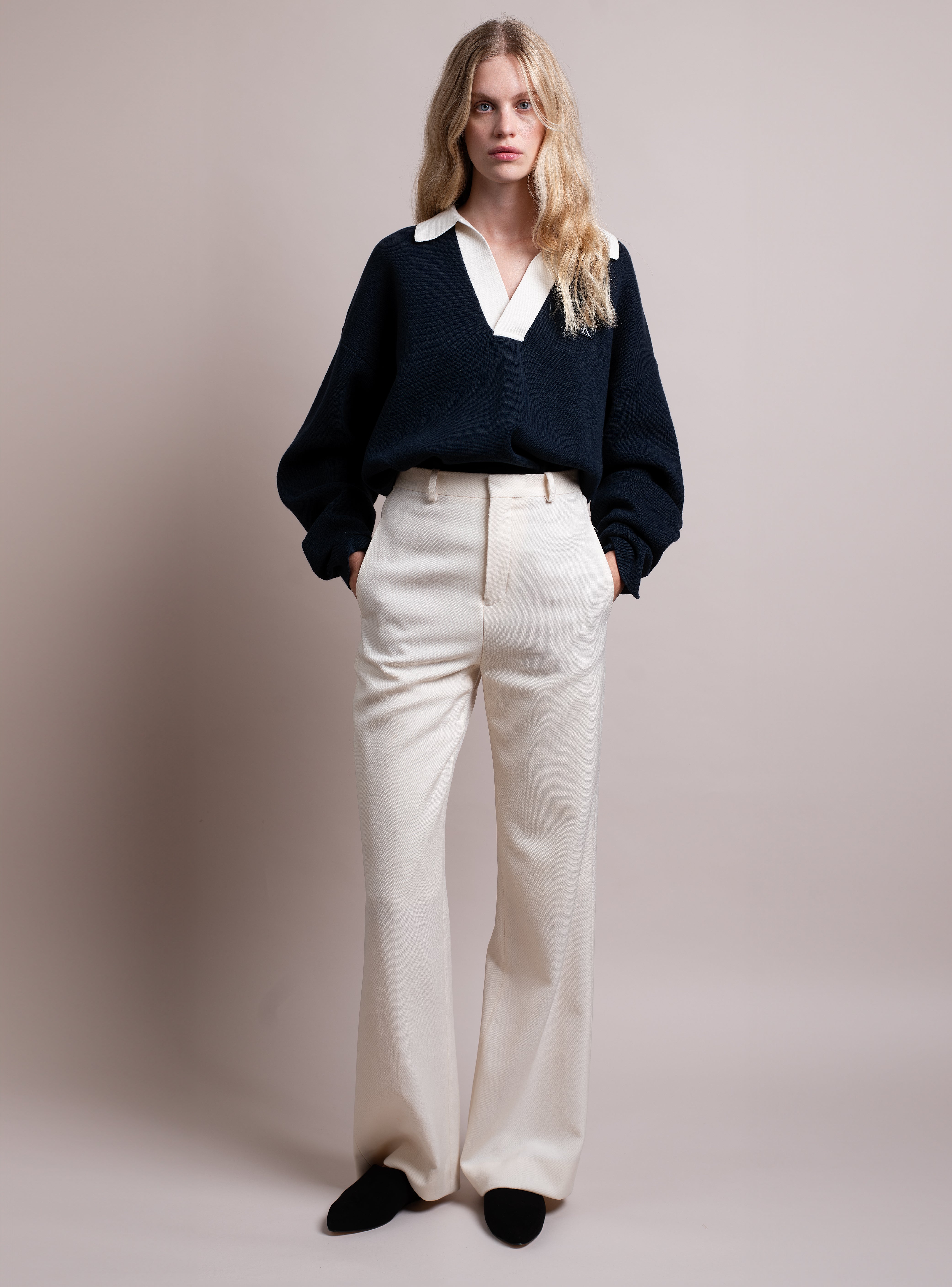 Woman wearing a navy sweater with a white collar, white pants, and black suede slippers with a pointy toe on a plain background
