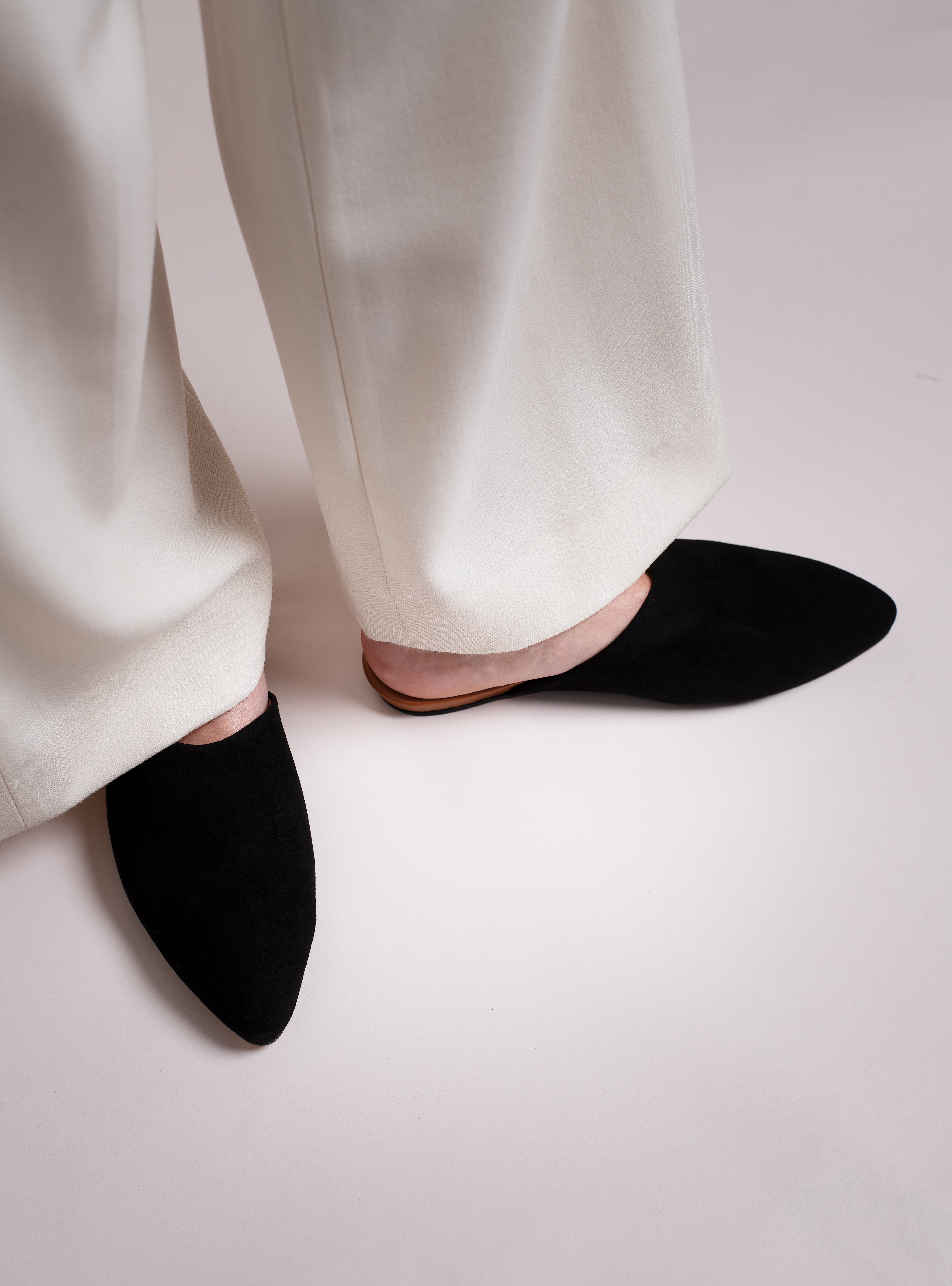 Woman wearing black suede pointed-toe slippers worn with white pants on a light background