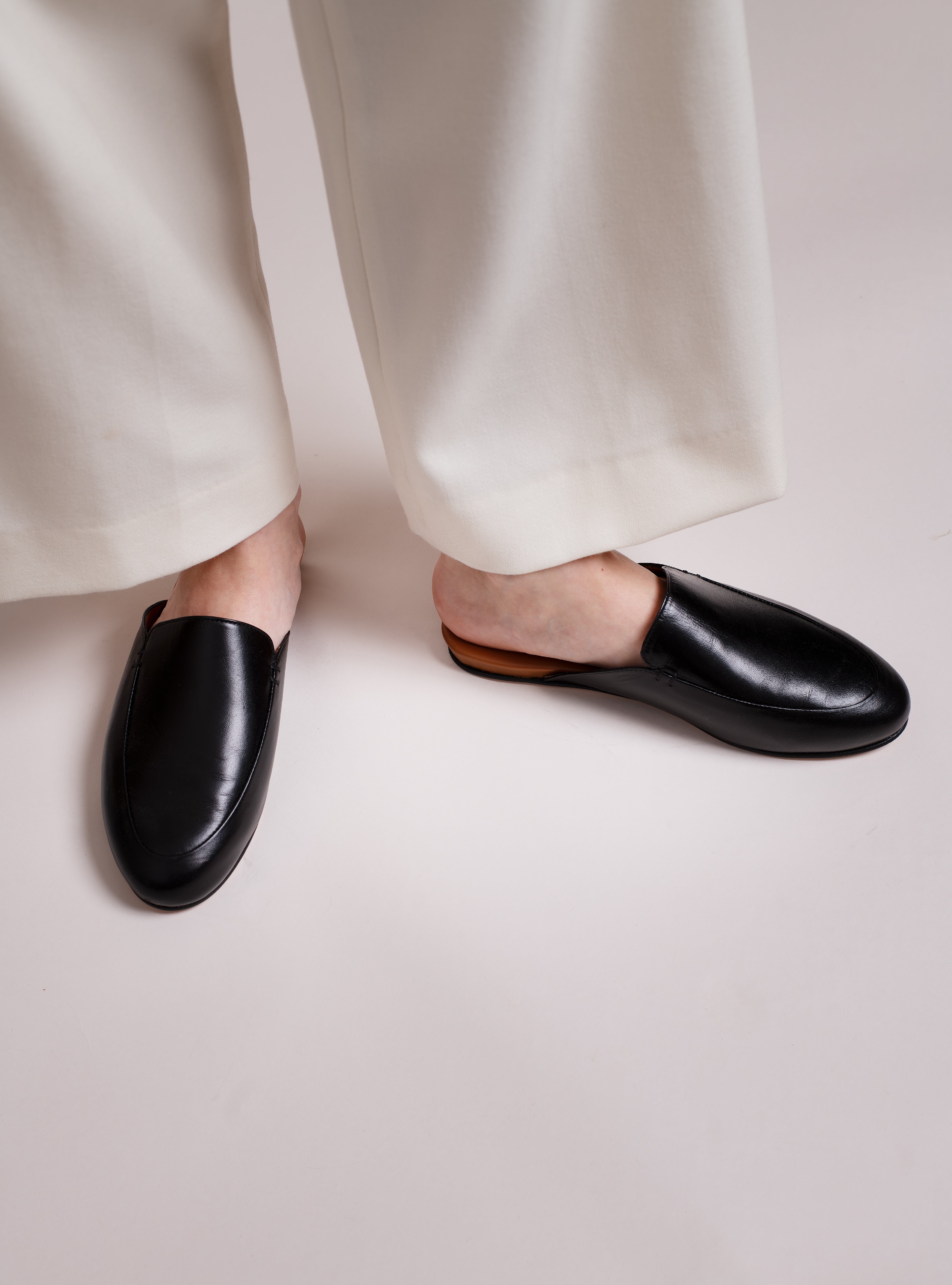 Woman wearing black leather slip-in loafers worn with white pants on a light gray background.