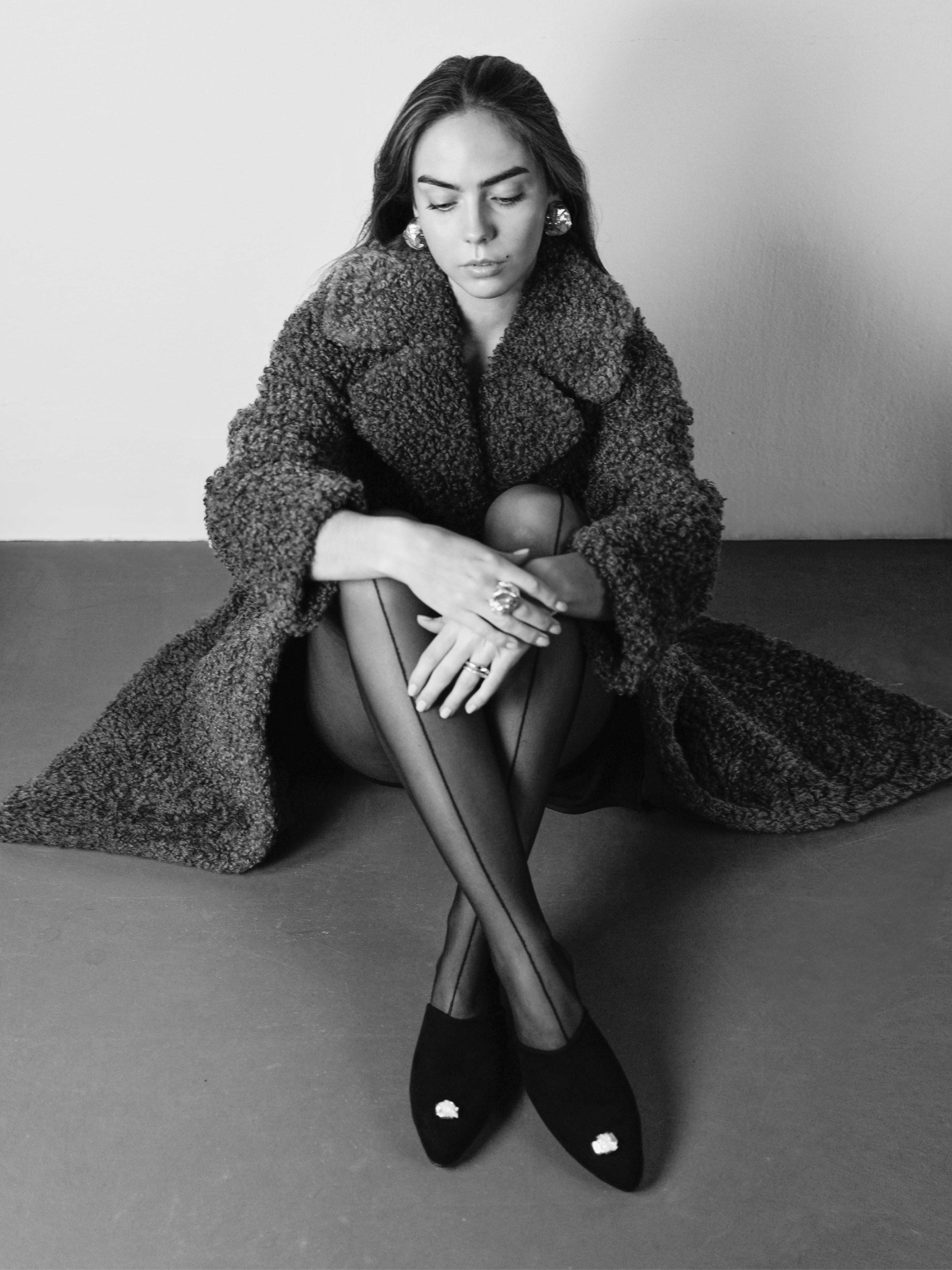Woman wearing a shearling coat and black suede slippers with a silver jewelry, sitting on a plain background.