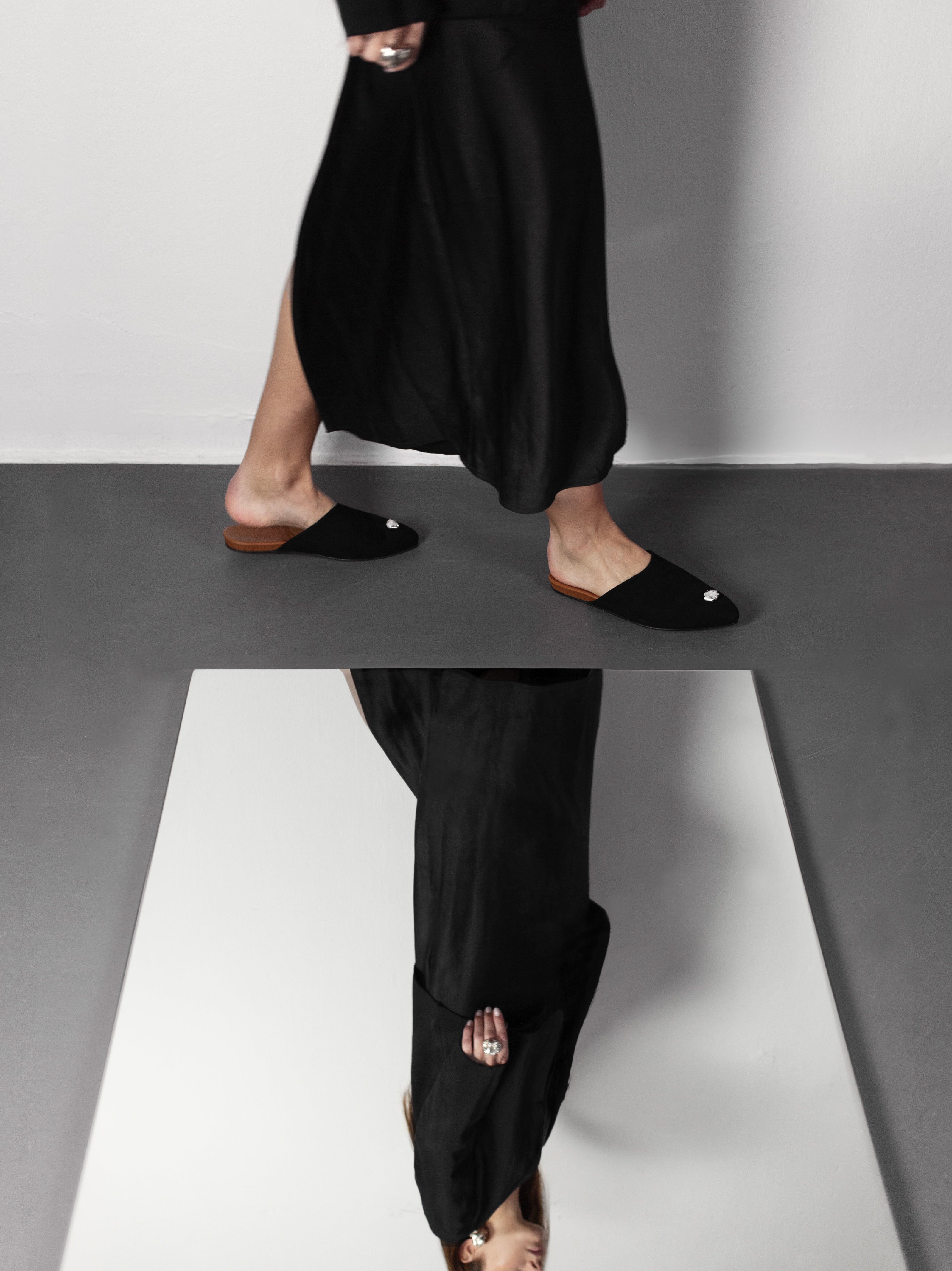 Woman wearing a black satin dress and black suede slippers with silver jewelry on a gray floor.