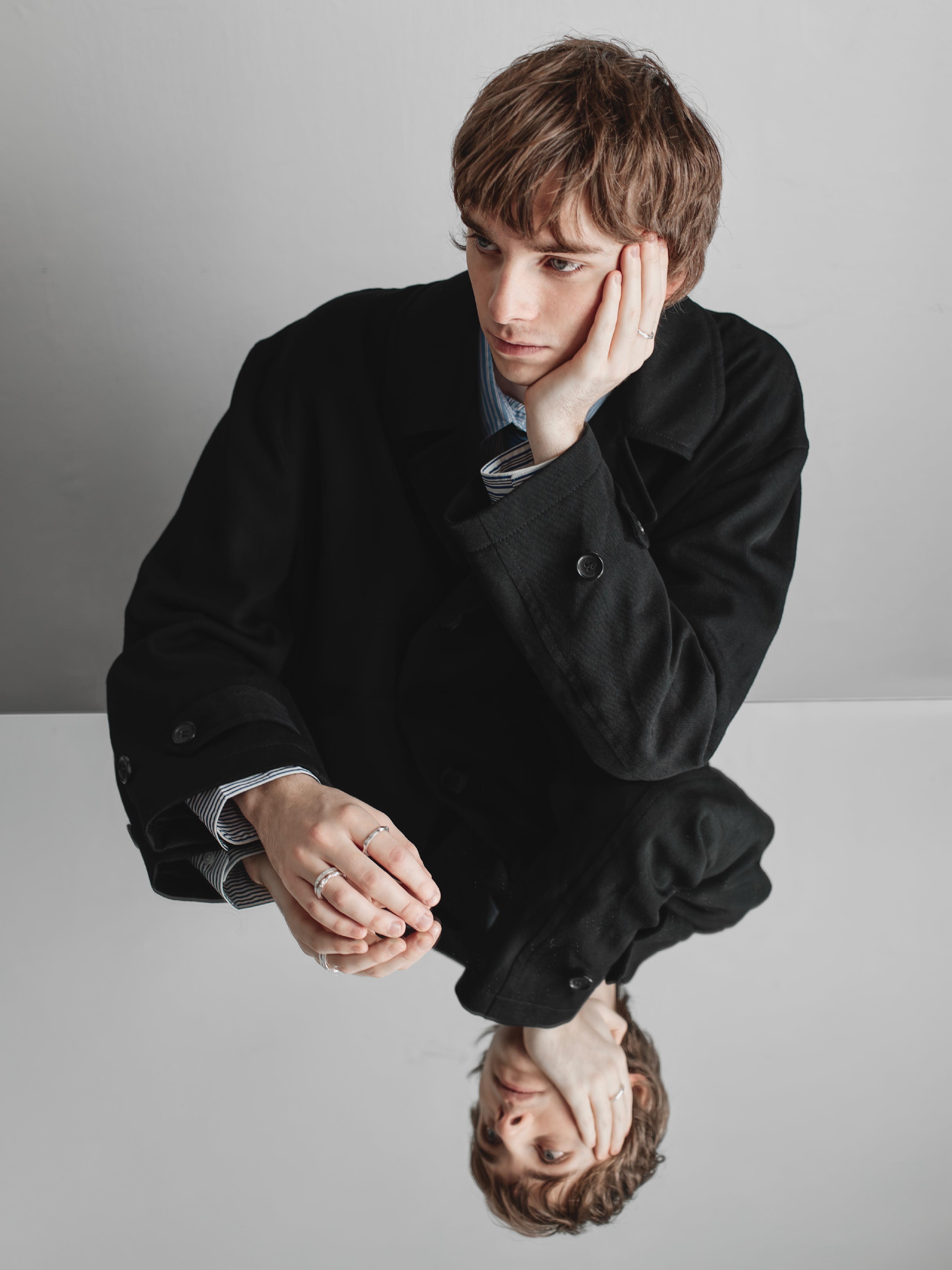 Man in a black coat sitting on a reflective surface with a neutral background