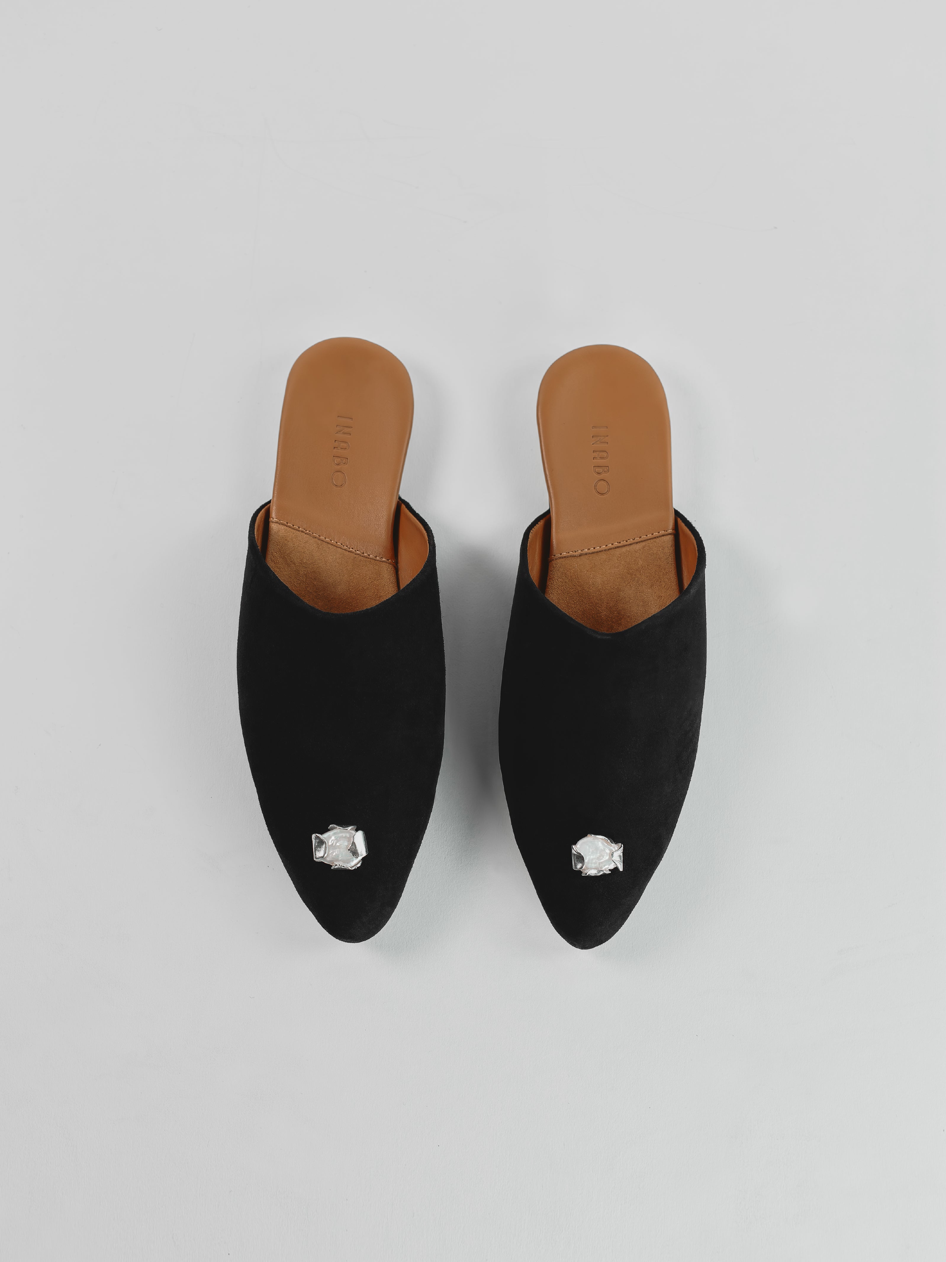Black pointed-toe slippers with brown insole and adorned with silver jewelry on a light gray background